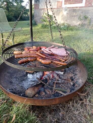 Verschiedene Würstchen und Fleischstücke werden auf einem runden Grill über einer offenen Feuerstelle im Freien auf dem grünen Rasen neben einem Gebäude gegrillt.