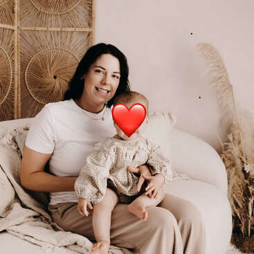 A woman with dark hair, wearing a white shirt, is sitting on a cozy sofa, holding a baby with a heart emoji covering their face. Pampas grass decorates the background.