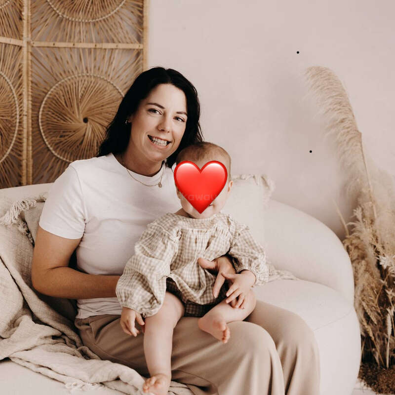 Una mujer de pelo oscuro, con camisa blanca, está sentada en un sofá acogedor, con un bebé en brazos y un emoji de corazón cubriéndoles la cara. La hierba de la Pampa decora el fondo.
