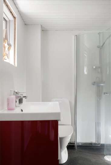 Esta imagem mostra um banheiro pequeno e moderno com paredes brancas, um lavatório vermelho, um vaso sanitário, uma cabine de chuveiro de vidro e uma janela com uma decoração em forma de estrela.