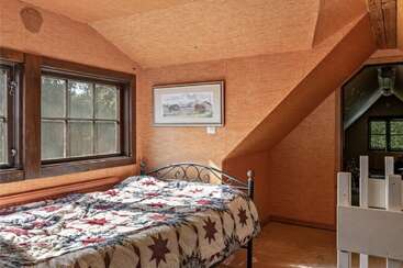 Este aconchegante quarto no sótão tem paredes inclinadas na cor laranja, uma janela com molduras de madeira, uma cama de metal preta, uma colcha com estampa de estrelas e uma pintura emoldurada com um cenário rural.