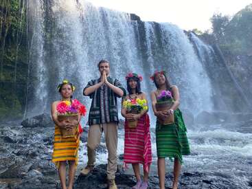 Cuatro personas vestidas con trajes tradicionales ante una gran cascada. Tres mujeres sostienen cestas de flores; el hombre está de pie con las palmas de las manos juntas, aparentando saludar o rezar.