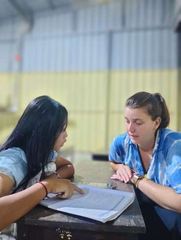 Una joven y una mujer se sientan a una mesa, concentradas en el papeleo. La chica señala el documento mientras la mujer escucha atentamente, en el interior.