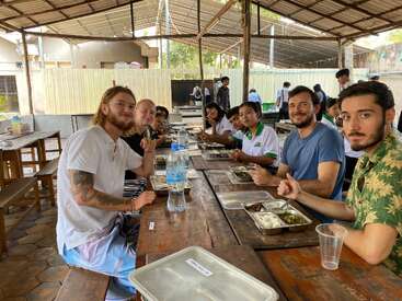 Un grupo de personas, entre las que hay estudiantes y adultos, están sentadas juntas en una larga mesa de madera en una cantina cubierta al aire libre, disfrutando de una comida y sonriendo.