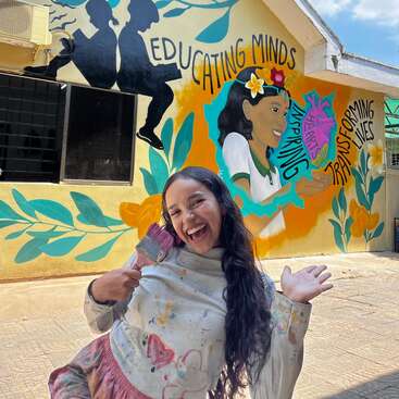 Una alegre mujer frente a un colorido mural pintado por ella misma, con mensajes positivos sobre educación, inspiración y transformación, con vibrantes flores y siluetas.