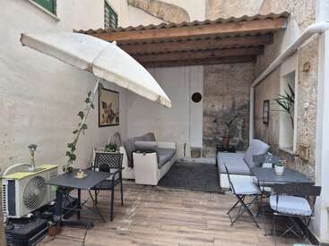 This cozy outdoor patio features a white umbrella, comfy sofas, potted plants, small tables with chairs, rug, and rustic stone walls covered by a wooden roof.