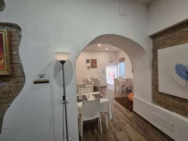 A cozy, modern restaurant interior with white walls, wooden floor, and archway. White tables and chairs are arranged neatly, complemented by wall art and soft lighting.