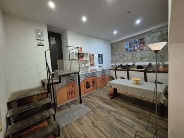 The image shows a small, cozy room with wooden flooring, a staircase, a table with a patterned tablecloth, a counter, shelving, and warm lighting.