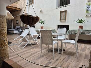 This image shows a cozy indoor courtyard with white chairs and tables, potted plants, a hanging planter, wooden flooring, and a "No Smoking" sign on the wall.