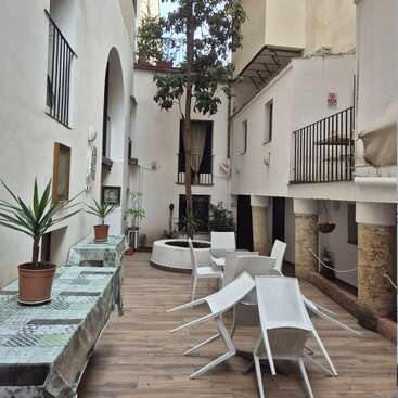 This image shows a narrow courtyard with white walls, potted plants on covered tables, a central tree, stacked white chairs, and surrounding balconies above. Peaceful atmosphere.