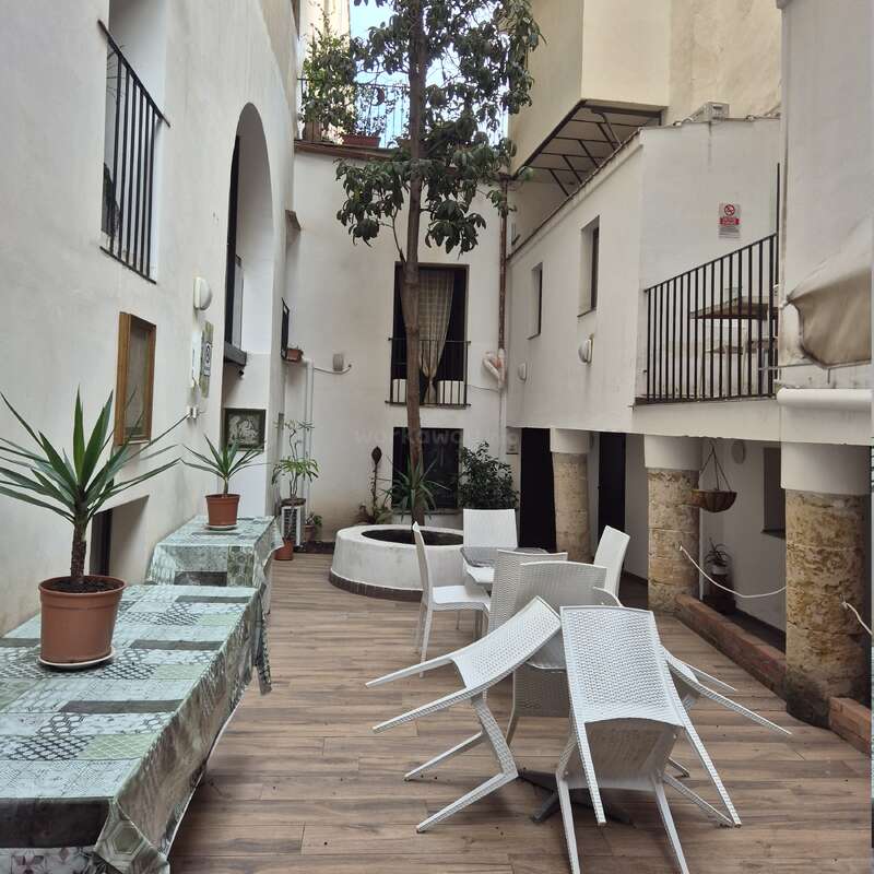 This image shows a narrow courtyard with white walls, potted plants on covered tables, a central tree, stacked white chairs, and surrounding balconies above. Peaceful atmosphere.