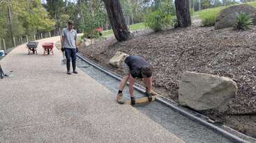 Duas pessoas estão trabalhando em um caminho dentro de um parque, espalhando e nivelando concreto. Árvores, pedras grandes e carrinhos de mão podem ser vistos no cenário natural ao redor.
