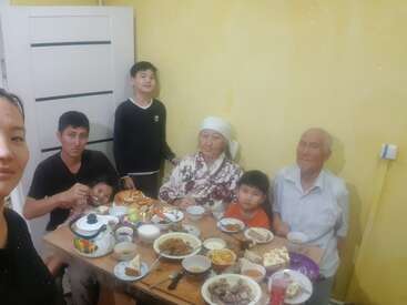 The image depicts a family of seven gathered around a table laden with an assortment of food, set against a yellow wall with a white door in the background.