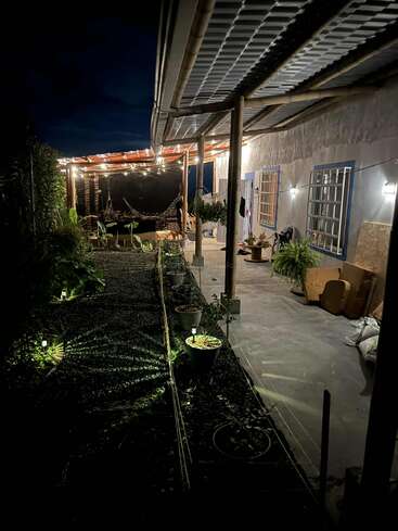 Eine gemütliche, rustikale Veranda bei Nacht, beleuchtet von Lichterketten und Gartenlampen. Topfpflanzen und Möbel säumen den Gehweg neben einem üppigen, eingezäunten Gartenbereich. Friedliche Atmosphäre.