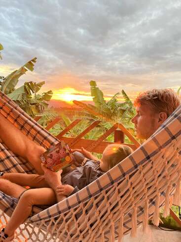 Ein Mann und ein Kind entspannen sich in einer Hängematte, lesen gemeinsam ein Buch und sind von tropischen Pflanzen umgeben, während ein wunderschöner Sonnenuntergang den Himmel in orangefarbenen Tönen färbt.