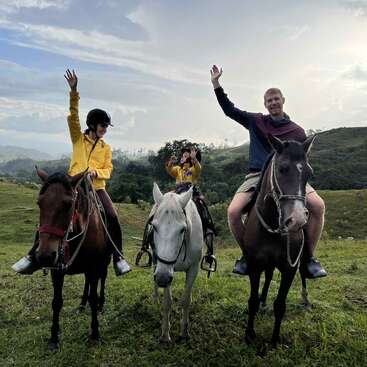 Drei Menschen sitzen auf Pferden im Freien, lächeln und winken. Sie tragen legere Kleidung und Helme, sind umgeben von grünen Hügeln und einem wolkenverhangenen blauen Himmel und genießen gemeinsam die Natur.