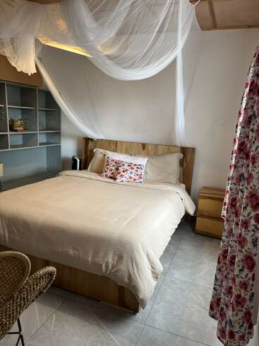 Ein gemütliches Schlafzimmer mit einem Holzbett, neutraler Bettwäsche, geblümten Kissen, einem Baldachin aus Moskitonetz, einem offenen Regal, einem Korbsessel, einem Nachttisch und einem geblümten Vorhang, der eine warme Atmosphäre schafft.