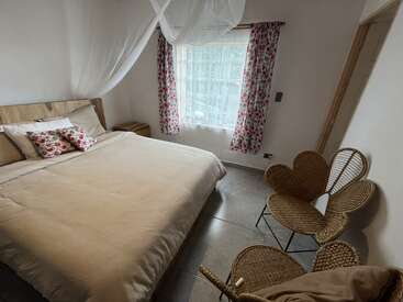 Este acogedor dormitorio cuenta con una cama doble con ropa de cama beige, almohadas de flores, cortinas de flores, sillas de mimbre, una mesa auxiliar, suelo de baldosas y mosquitera.