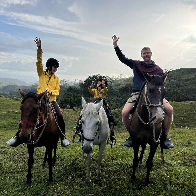 Drei Menschen sitzen auf Pferden im Freien, lächeln und winken. Sie tragen legere Kleidung und Helme, sind umgeben von grünen Hügeln und einem wolkenverhangenen blauen Himmel und genießen gemeinsam die Natur.