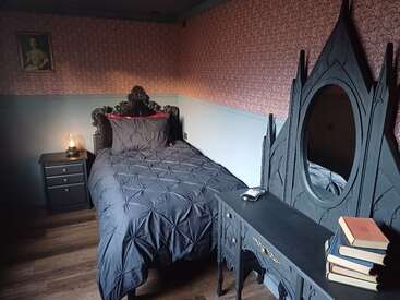 Ein Schlafzimmer im Gothic-Stil mit einem geschnitzten Bett, dunkler Bettwäsche, einem verzierten Spiegel, einem Beistelltisch mit einer Lampe, gestapelten Büchern und einer Vintage-Tapete schafft eine stimmungsvolle Atmosphäre.