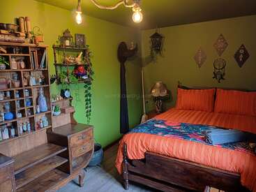 In diesem gemütlichen Schlafzimmer sorgen grüne Wände, Holzmöbel, eine eklektische Einrichtung, ein Doppelbett mit orangefarbener Bettwäsche, mystische Gegenstände, hängende Pflanzen und eine warme Raumbeleuchtung für eine skurrile Atmosphäre.