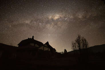 Ein Haus steht in der Silhouette unter einem atemberaubenden, sternenübersäten Nachthimmel. Die Milchstraße leuchtet hell auf und erhellt die friedliche, ruhige Landschaft mit himmlischer Schönheit.