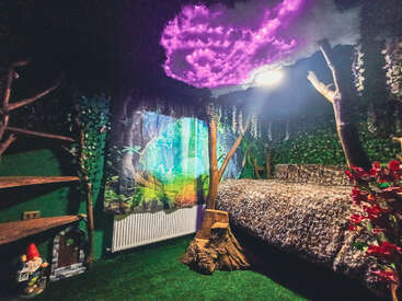 Dieses bezaubernde Schlafzimmer hat ein magisches Waldthema mit künstlichen Bäumen, belaubten Wänden, einer leuchtenden lila Wolkendecke, Walddekoration und einem gemütlichen, getarnten Bett.