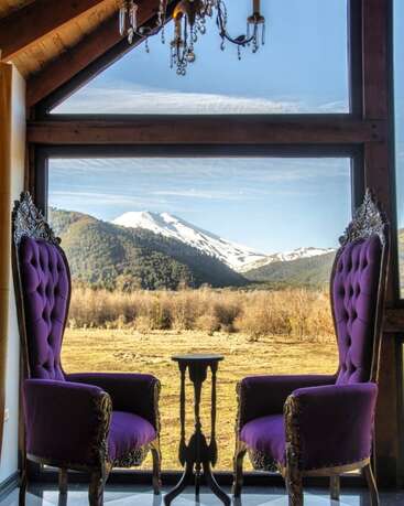 Zwei verzierte lilafarbene Sessel stehen sich an einem kleinen Tisch gegenüber, der vor einem großen Fenster steht, das den Blick auf eine atemberaubende schneebedeckte Berglandschaft und den blauen Himmel freigibt.