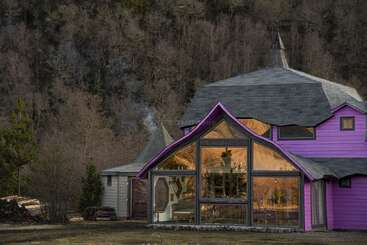 Ein skurriles lila Haus mit großen Glasfenstern steht auf einer Waldlichtung. Das gemütliche Innere leuchtet warm und steht im Kontrast zu den kahlen Bäumen und der gedämpften Landschaft.