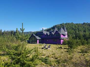 A vibrant purple house with pointed roofs sits in a lush green field, surrounded by trees and hills under a clear blue sky with Adirondack chairs nearby.