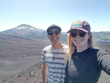 Das Bild zeigt einen Mann und eine Frau, die zusammen vor einer Berglandschaft stehen. Beide tragen Hüte und Sonnenbrillen, wobei der Arm der Frau in Richtung Kamera ausgestreckt ist.