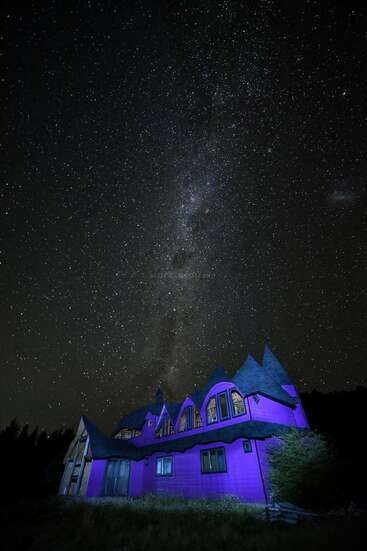 Das Bild zeigt ein majestätisches lilafarbenes Haus mit einem dunklen Dach vor einem sternenklaren Nachthimmel, der ein Gefühl von Gelassenheit und Verzauberung ausstrahlt.