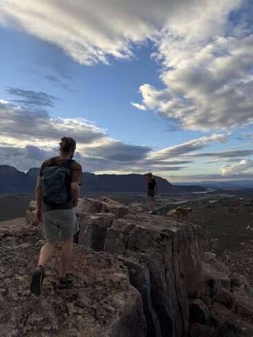 Deux personnes et un chien se tiennent au sommet de rochers escarpés, contemplant un vaste paysage sous un ciel orageux, en randonnée au-dessus d'un terrain désertique, avec au loin un magnifique panorama montagneux à perte de vue.