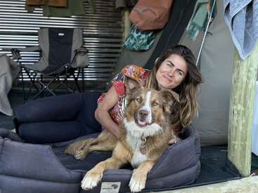 Une femme aux cheveux longs est allongée à côté d'un chien brun et blanc sur un panier pour chien ; tous deux se détendent dans une tente ou une cabane confortable.