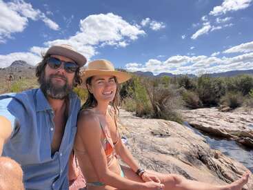 Un couple profite d'une journée ensoleillée en plein air, assis sur un terrain rocheux près d'un ruisseau. Ils sourient, vêtus de tenues d'été et coiffés de chapeaux, avec de magnifiques montagnes en arrière-plan.