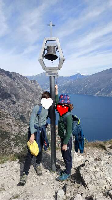 Zwei Kinder stehen neben einer Metallglocke auf einem Berggipfel. Sie überblicken einen wunderschönen blauen See, umgeben von schroffen Felsen und einem weiten Himmel. Die Gesichter sind unkenntlich gemacht.
