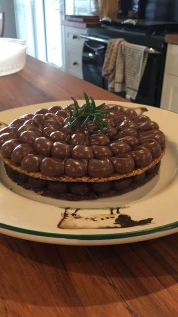 Das Bild zeigt ein Schokoladendessert auf einem weißen Teller mit grünem Rand, der auf einem Holztisch in einer Küche steht.