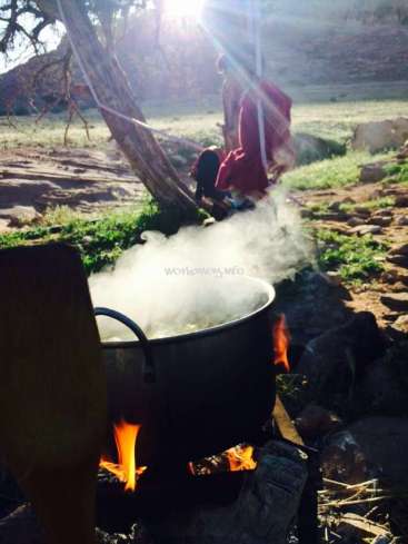 Das Bild zeigt einen großen Topf mit kochendem Wasser über einem Feuer, in dessen Nähe eine Person in einem roten Gewand vor einer natürlichen Kulisse steht.