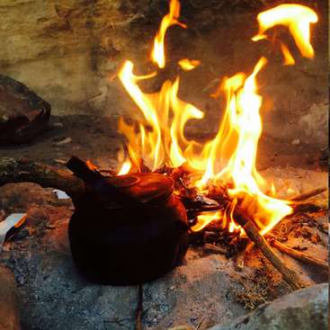 Das Bild zeigt einen Kessel, der auf einem Felsen neben einem Feuer sitzt und dessen Flammen aus dem Brennholz aufsteigen. Der Kessel ist schwarz und scheint aus Metall zu sein.