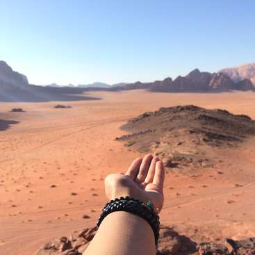 Das Bild zeigt eine Hand mit Armbändern vor einer weiten Wüstenlandschaft mit Sanddünen, Felsen und fernen Bergen unter einem klaren blauen Himmel.