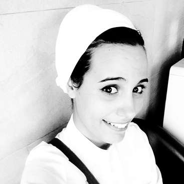 Das Bild ist ein schwarz-weißes Selfie einer Frau, die eine weiße Kopfbedeckung und ein weißes Hemd trägt, mit einem schwarzen Riemen über der Schulter, vor einem hellen Wandhintergrund.