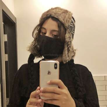 Das Bild zeigt eine Person, die ein Selfie in einem Spiegel macht, einen schwarzen Mantel und Hut trägt und ein weißes iPhone in der Hand hält, vor einem weißen Wandhintergrund.