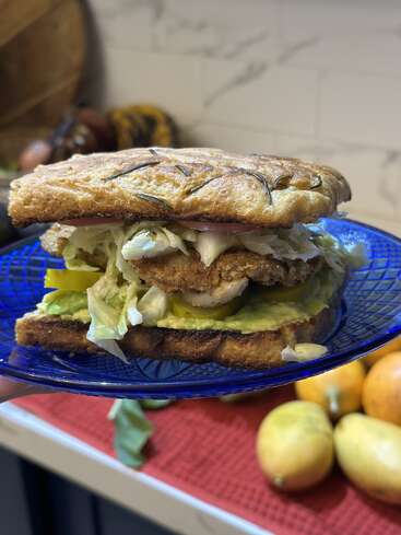 A delicious sandwich with crispy bread, fried patty, fresh lettuce, tomato, avocado spread, and pickles sits on a blue plate in a cozy kitchen.