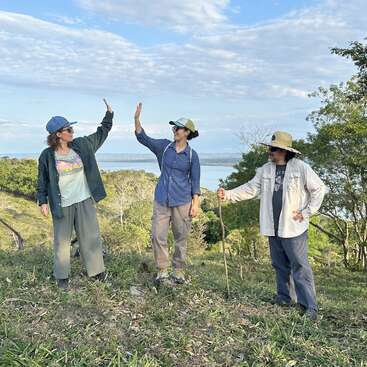 Three people stand outdoors on a grassy hill under a blue sky, two giving a high-five, all wearing hats, enjoying nature and each other's company.