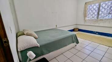 This is a simple bedroom with white tiled floors, a green bedspread, pillows, a small lamp, a window with lace curtains, and minimal decoration.