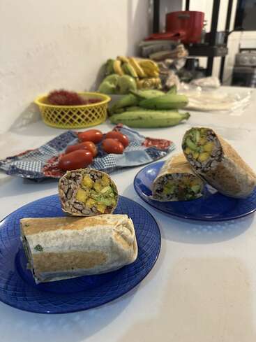 Two burritos on blue plates are cut open to show rice, beans, and vegetables. In the background are tomatoes, plantains, bananas, and a yellow fruit basket.
