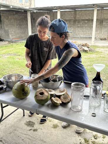 Two people are preparing fresh coconuts at an outdoor table. There are bowls, jars, and coconut shells, suggesting they are extracting or enjoying coconut water together.