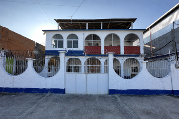 This image shows a two-story white building with arched windows and gates, red brick accents, barbed wire, and a blue trim, surrounded by a concrete yard.
