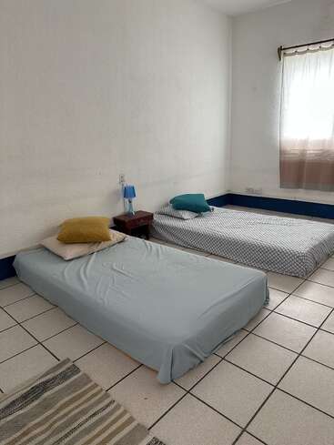 Das Bild zeigt ein einfaches, minimalistisches Schlafzimmer mit zwei niedrigen Betten, einem Nachttisch, einer kleinen blauen Lampe, Fliesenboden, Teppich und einem Fenster mit transparenten Vorhängen.