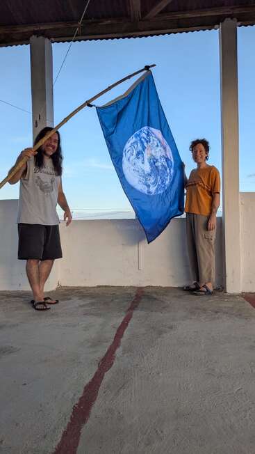 Zwei Leute lächeln, während sie eine blaue Flagge mit einem Bild der Erde hochhalten. Sie stehen auf einem Dach unter einem einfachen Unterstand und genießen den klaren Himmel.